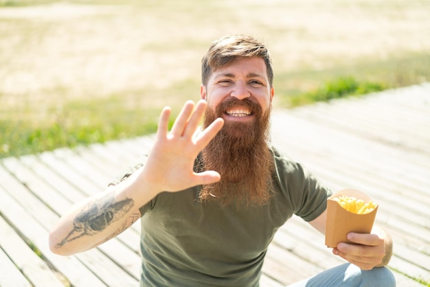Rudy mężczyzna z brodą trzymający smażone frytki na zewnątrz pozdrawiając ręką z radosnym wyrazem twarzy
