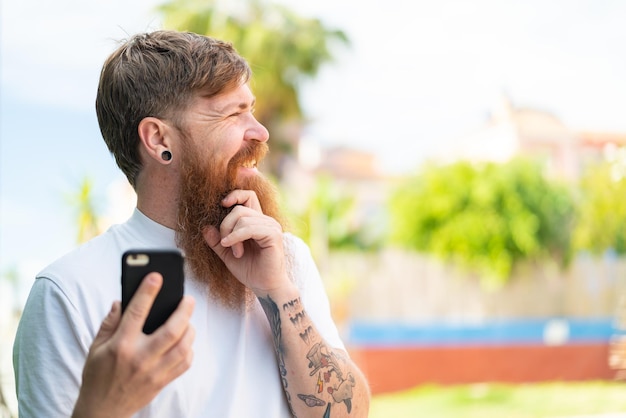 Rudy mężczyzna z brodą za pomocą telefonu komórkowego na zewnątrz, myśląc o pomyśle i patrząc z boku