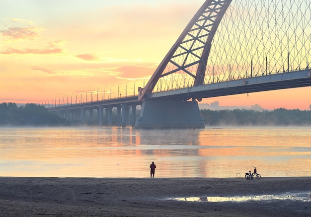 Rybak na plaży. Samochód Bugrinskij most przez Wielką Syberyjską rzekę Ob w porannej mgle