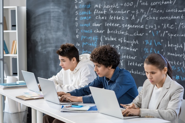 Rząd trzech współczesnych poważnych studentów pracujących przed laptopami, siedząc przy biurku