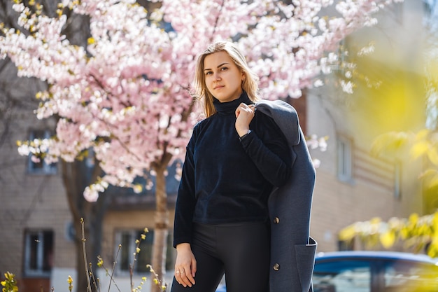 Sakura Rozgałęzia Się Z Kwiatami Na Drzewie Na Ulicach Miasta. Szczęśliwa Kobieta Dziewczyna W Szarej Palecie Spaceruje Aleją Z Kwitnącą Sakura. Wspaniała Fantazyjna Dziewczyna Na Zewnątrz. Kwitnące Drzewo Sakura.