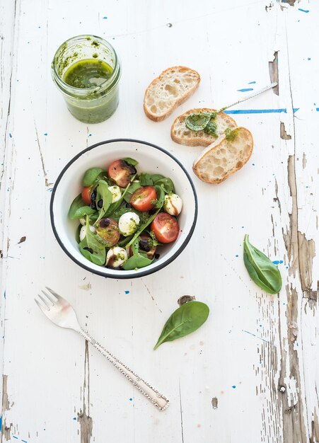 Zdjęcie sałatka caprese chiabatta plastry cherrytomatoes baby szpinak i mozzarella w metalowej misce z dressingiem pesto na rustykalnym białym drewnianym tle