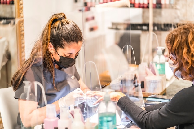 Salon manicure i pedicure, koronawirus, COVID-19, dystans społeczny. Ponowne otwarcie z powodu pandemii, środki bezpieczeństwa. Szczegółowo pracownica za ekranem ochronnym