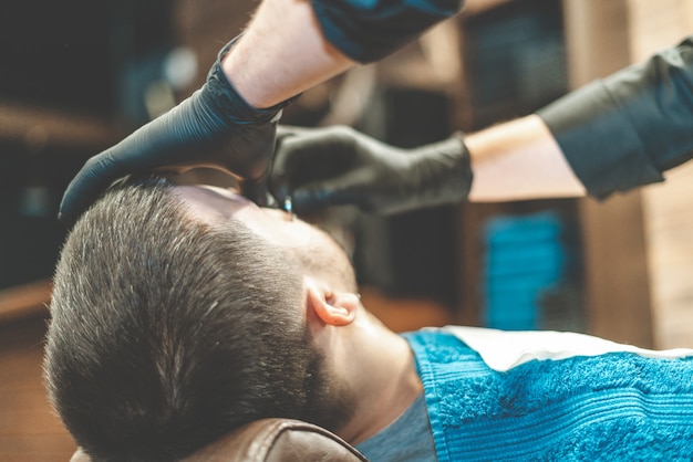Salon piękności dla mężczyzn. Golenie brody w zakładzie fryzjerskim. Fryzjer przycina brodę brzytwą i maszynką do strzyżenia. bliska Brutalne fryzury. Sprzęt fryzjerski. Selektywne ustawianie ostrości.
