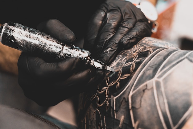 Salon tatuażu. Mistrz tatuażu tatuuje mężczyznę na ramieniu. Maszynka do tatuażu, bezpieczeństwo i higiena w pracy. Makro, przyciemniane, tatuażysta