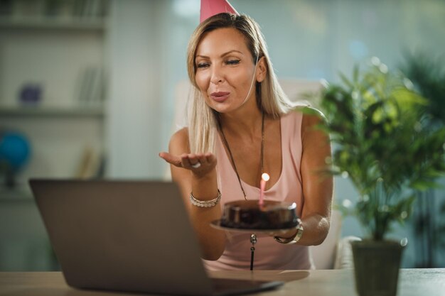 Sama atrakcyjna kobieta obchodzi urodziny w domu podczas pandemicznej izolacji i prowadzi wideorozmowę z przyjaciółmi. Ona trzyma tort urodzinowy z zapalonymi świeczkami.