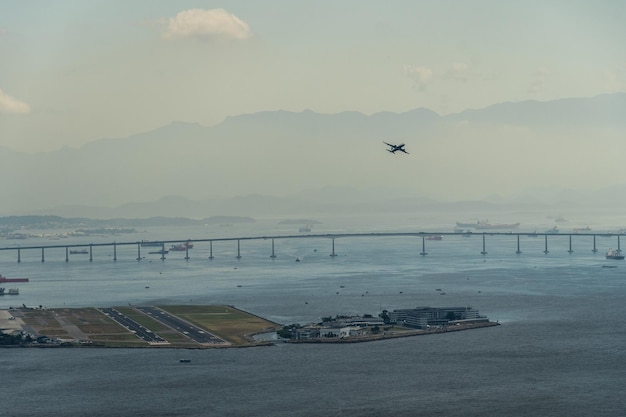 Samolot startujący z pasa startowego lotniska w Rio de Janeiro Brazylia Rio Niteroi Most górski i statki w tle Świt z lekką mgłą