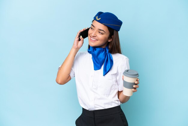 Samolot stewardessa kaukaska kobieta na białym tle na niebieskim tle trzymająca kawę na wynos i telefon komórkowy