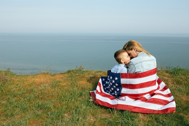 Samotna Matka Z Synem W Dzień Niepodległości Usa. Kobieta I Jej Dziecko Chodzą Z Flagą Usa Na Wybrzeżu Oceanu