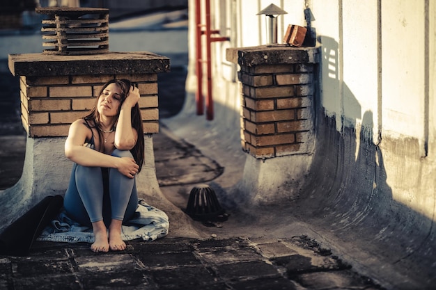 Samotna młoda kobieta w depresji siedzi z kolanami w objęciach, zagubiona w złych pesymistycznych myślach i przygotowująca się do treningu na tarasie na dachu.