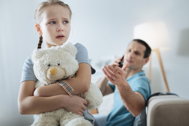 Samotna niebieskooka jasnowłosa dziewczynka trzymająca zabawkę i płacząca, podczas gdy jej ojciec rozmawia przez telefon