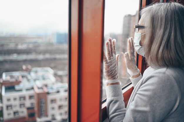 Samotna starsza starsza smutna kobieta w masce medycznej i rękawiczkach, patrząc na okno, pozostaje izolacją w domu dla własnej kwarantanny. Kwarantanna domowa, zapobieganie COVID-19, Sytuacja epidemii koronawirusa