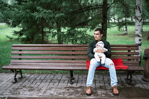 Samotny tata w zielonej kurtce siedzi na ławce i przytulanie swojego dziecka w parku, koncepcja ojcostwa