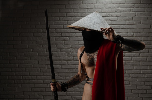 Samuraj w czerwonej pelerynie i azjatyckim trójkątnym kapeluszu z tatuażami i mieczem katana w dłoniach na tle ceglanego muru