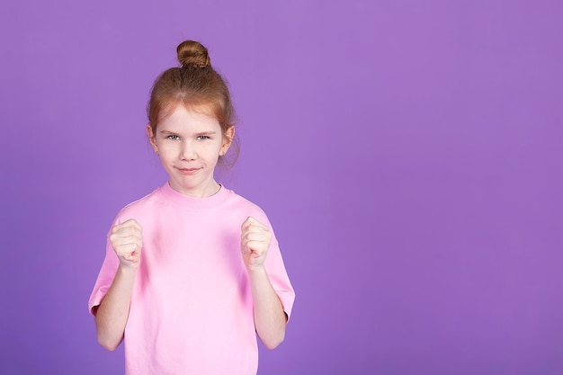 Sceptyczna dziewczynka ubrana w różową koszulkę i dżinsy stoi na fioletowym tle studia trzymając ręce na pasku