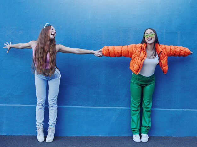 Zdjęcie Ściana przyjaciół i miasto dla mody z dziewczyną trzymającą się za ręce i na zewnątrz dla śmiesznej komicznej twarzy lub szczęśliwych razem kobiet gen z metra i niebieskiego tła dla modnych ubrań trampki lub miejskiej przygody