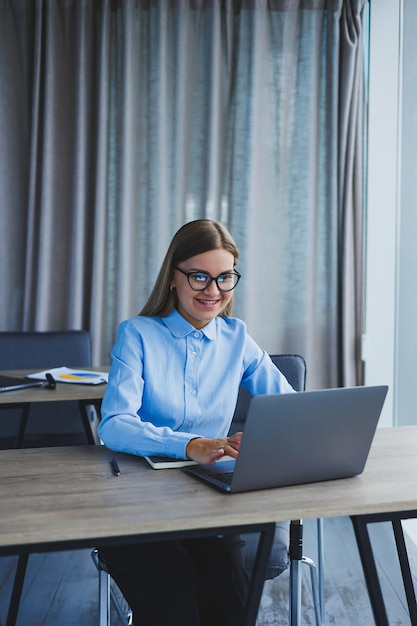Sekretarka sprawdza harmonogram Wesoła młoda piękna kobieta w okularach z uśmiechem korzysta z laptopa siedząc w miejscu pracy