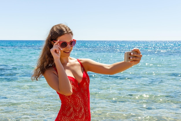 Seksowna dziewczyna na plaży ze zdziwionym wyrazem twarzy patrząca na telefon i robiąca selfie