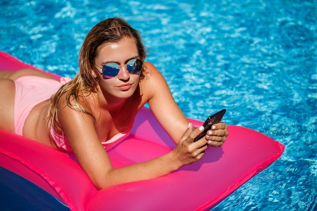 Seksowna kobieta z telefonem w stroju kąpielowym leży na różowym dmuchanym materacu w basenie. Zrelaksuj się przy basenie w upalny, słoneczny dzień. Koncepcja wakacji