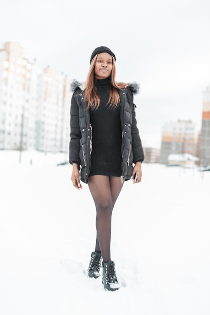 Seksowna ładna Młoda Afrykańska Kobieta W Modne Zimowe Ciepłe Ubrania Z Kurtką, Czapką, Rajstopami I Butami Spaceruje Po Ulicy Ze śniegiem Na Tle Domów