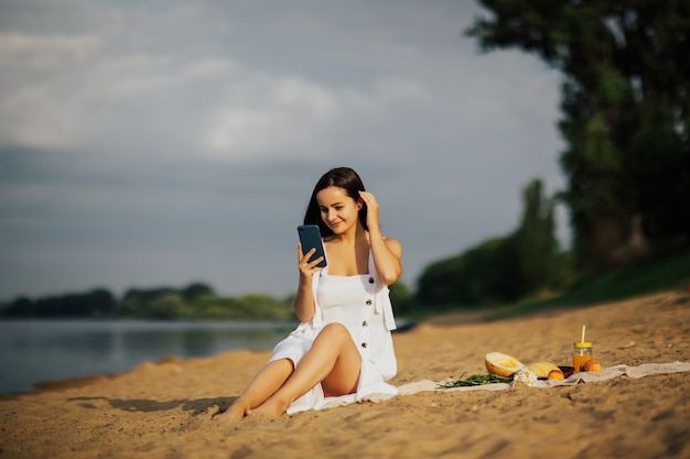 Seksowna młoda kobieta na plaży przy użyciu smartfona. Używała telefonu komórkowego podczas letniego pikniku na plaży, pijąc soki i jedząc owoce.