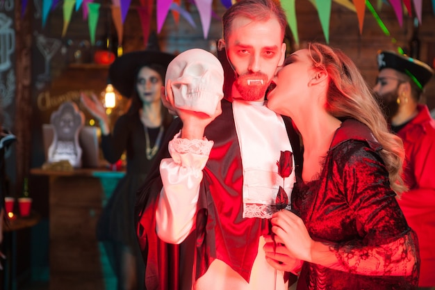 Zdjęcie seksowna młoda kobieta przebrana za wampira gryzącego szyję mężczyzny przebrana za drakulę na halloween. dracula trzyma ludzką czaszkę.