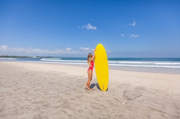 Seksowna młoda kobieta z długimi włosami w czerwonym swimsuit z deską surfingową samotnie na plaży