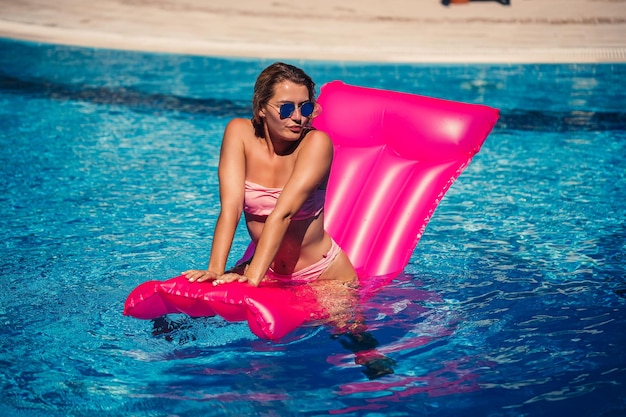 Seksowna modelka w okularach przeciwsłonecznych odpoczywa i opala się na materacu w basenie Kobieta w różowym stroju kąpielowym bikini unosząca się na dmuchanym różowym materacu z filtrem i filtrem przeciwsłonecznym