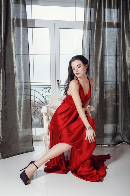 Seksowny glamour kobieta z czarnymi włosami w eleganckiej czerwonej sukience, siedząc na fotelu w luksusowej sypialni
