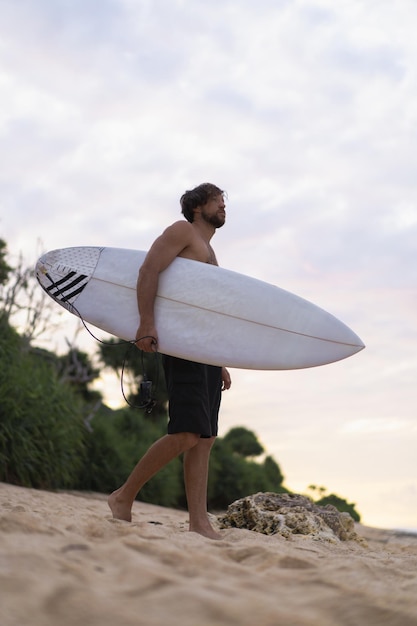 Seksowny surfer surfing człowiek z deską surfingową. Przystojny młody sportowiec mężczyzna trzyma deskę surfingową z mokrymi włosami na wakacje sport lato plaża. Sportowy cel podróży. Styl życia surfingu.