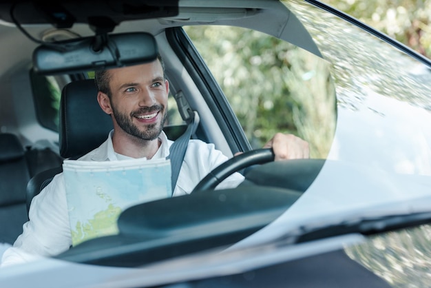 Selektywne skupienie brodatego i szczęśliwego mężczyzny prowadzącego samochód i patrzącego na mapę