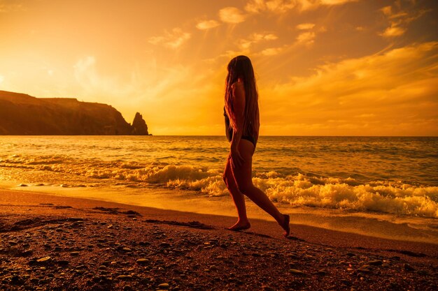 Selektywne skupienie szczęśliwa beztroska zmysłowa kobieta z długimi włosami w czarnym stroju kąpielowym pozuje na plaży o zachodzie słońca