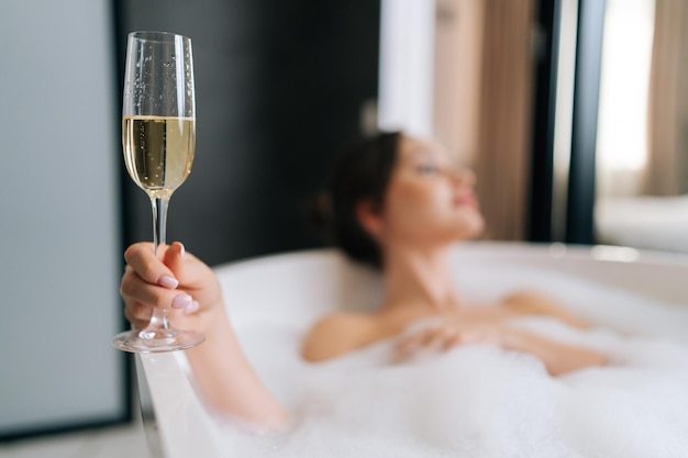 Selektywny fokus strzał zbliżenie młodej kobiety leżącej w wannie z pianką, trzymającej szklanego szampana, ciesząc się napojem alkoholowym podczas luksusowego wypoczynku na weekend