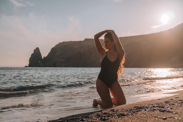 Selektywny fokus Szczęśliwa beztroska zmysłowa kobieta z długimi włosami w czarnym stroju kąpielowym pozowanie na plaży o zachodzie słońca Sylwetka młodej pięknej figlarnej pozytywnej kobiety na zewnątrz Letnie wakacje i koncepcja podróży