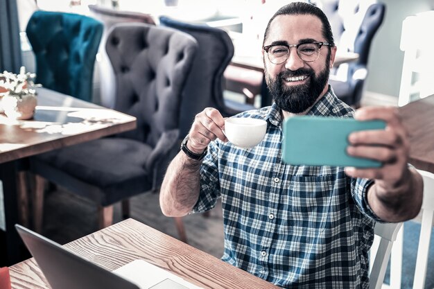 Selfie i kawa. Rozpromieniony brodaty ciemnowłosy mężczyzna czuje się zrelaksowany robiąc selfie i pijąc kawę