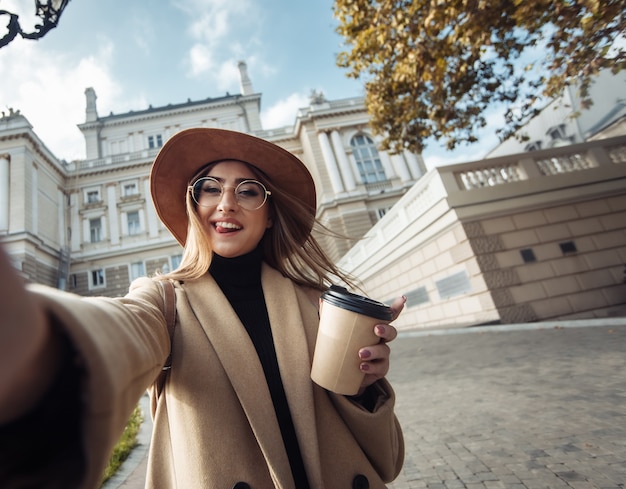 Selfie portret młodej stylowej turystki kobiety ubranej w płaszcz i kapelusz na tle europejskiej architektury miejskiej. Koncepcja wakacji i turystyki