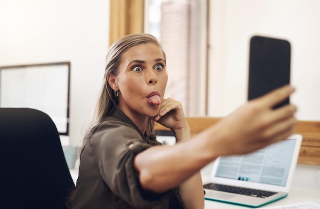 Selfie telefonem z zabawną biznesową kobietą bawiącą się głupkowatą i żartującą podczas pracy w biurze Młoda kobieta wystawiająca język i robiąca minę w pracy, czując się beztroska