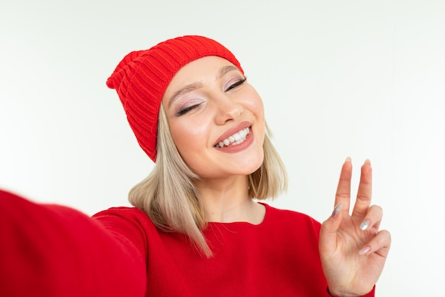 Selfie uśmiechnięta blondynki dziewczyna w czerwonym kapeluszu i pulowerze na białym tle