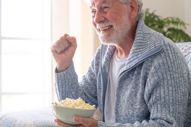 Senior 70s mężczyzna siedzący na kanapie jedzący popcorn wypoczynek i ludzie koncepcja szczęśliwy brodaty starszy mężczyzna relaksuje się w domu patrząc na telewizor