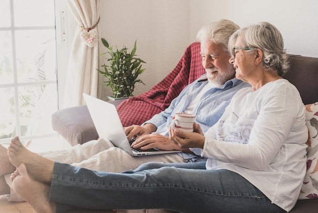 Senior kaukaski para siedzi w domu na kanapie za pomocą laptopa Dwóch starszych emerytów korzystających z wolnego czasu i urządzeń technologicznych