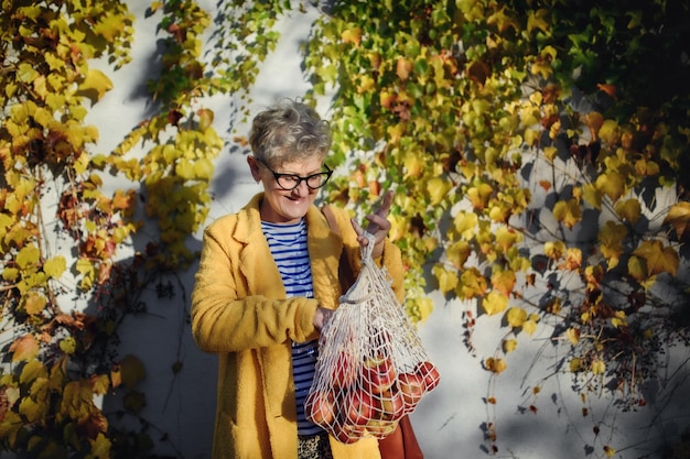 Senior kobieta stojąca na zewnątrz na kolorowym naturalnym tle jesieni, trzymając zakupy w torbie wielokrotnego użytku.
