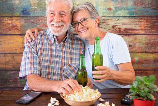 Senior para zabawy oglądając mecz piłki nożnej w telewizji, pijąc piwo i jedząc popcorn. Drewniane tło