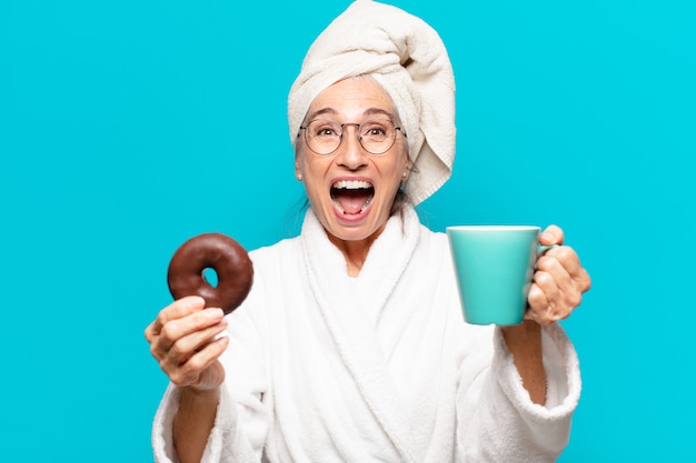 Senior pretty woman po prysznicem i nosząc szlafrok i jedząc śniadanie z kawą i pączkiem