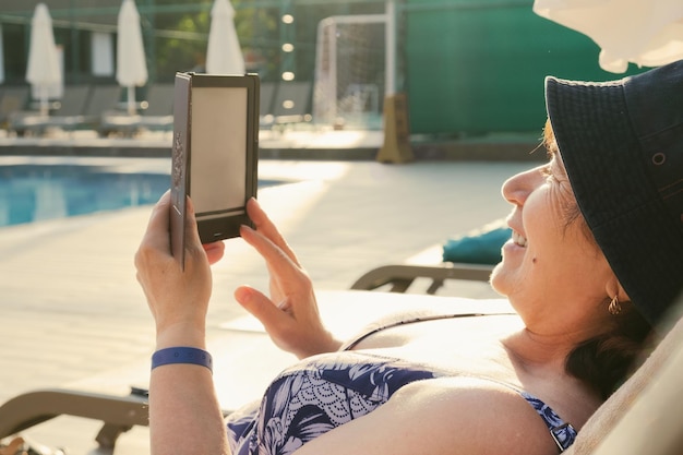 Seniorka w stroju kąpielowym na leżaku przy basenie czyta na urządzeniu elektronicznym