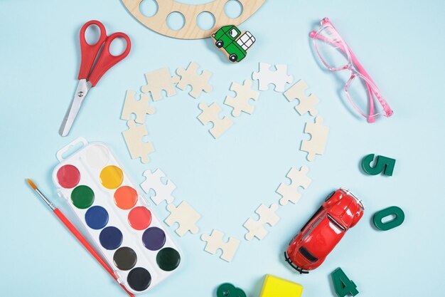 Serce z drewnianych puzzli i różnych przyborów szkolnych na niebieskim tle, farb, okularów dla dzieci i zabawek edukacyjnych, uczące małe dzieci i rozwijające wyobraźnię