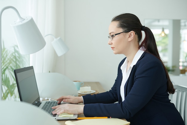 Seroius młoda kobieta przedsiębiorca w okularach siedzi przy stoliku w kawiarni i odpowiada na e-maile na laptopie