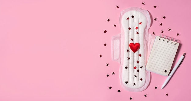 Serwetka sanitarna z czerwonymi płytkami i notatnikiem na różowym tle Okres cyklu miesiączkowego