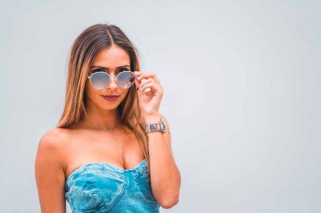Sesja Lifestyle, młoda kaukaska brunetka w niebieskiej dżinsowej sukni i okularach przeciwsłonecznych na prostym szarym tle