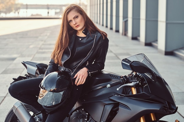 Sexy biker dziewczyna ubrana w czarną skórzaną kurtkę pozowanie na jej superbike poza budynkiem.