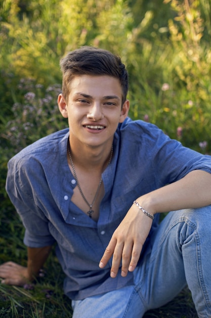 Sexy teen facet siedzi na trawie w przyrodzie w koszuli i dżinsach boso i patrząc w kamerę